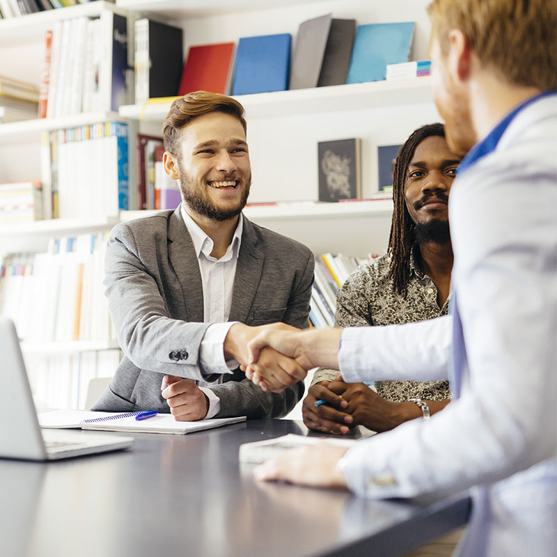 picture of two businessmen shaking hands with another business person sitting at the table
